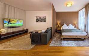 Een hotelkamer met een houten vloer, een bed met wit beddengoed, een bank met een plaid en een televisie die aan de muur is bevestigd, met daarboven een schilderij.