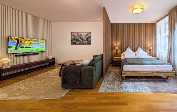 A hotel room with a wooden floor, a bed with white bedding, a sofa with a throw blanket, a television mounted on the wall, and a painting above the sofa.