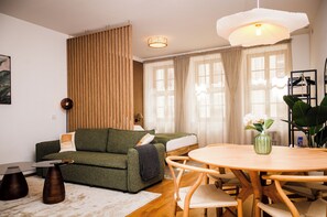 A modern living room with a green sofa, a wooden dining table with chairs, a hanging light fixture, and a large window with curtains.
