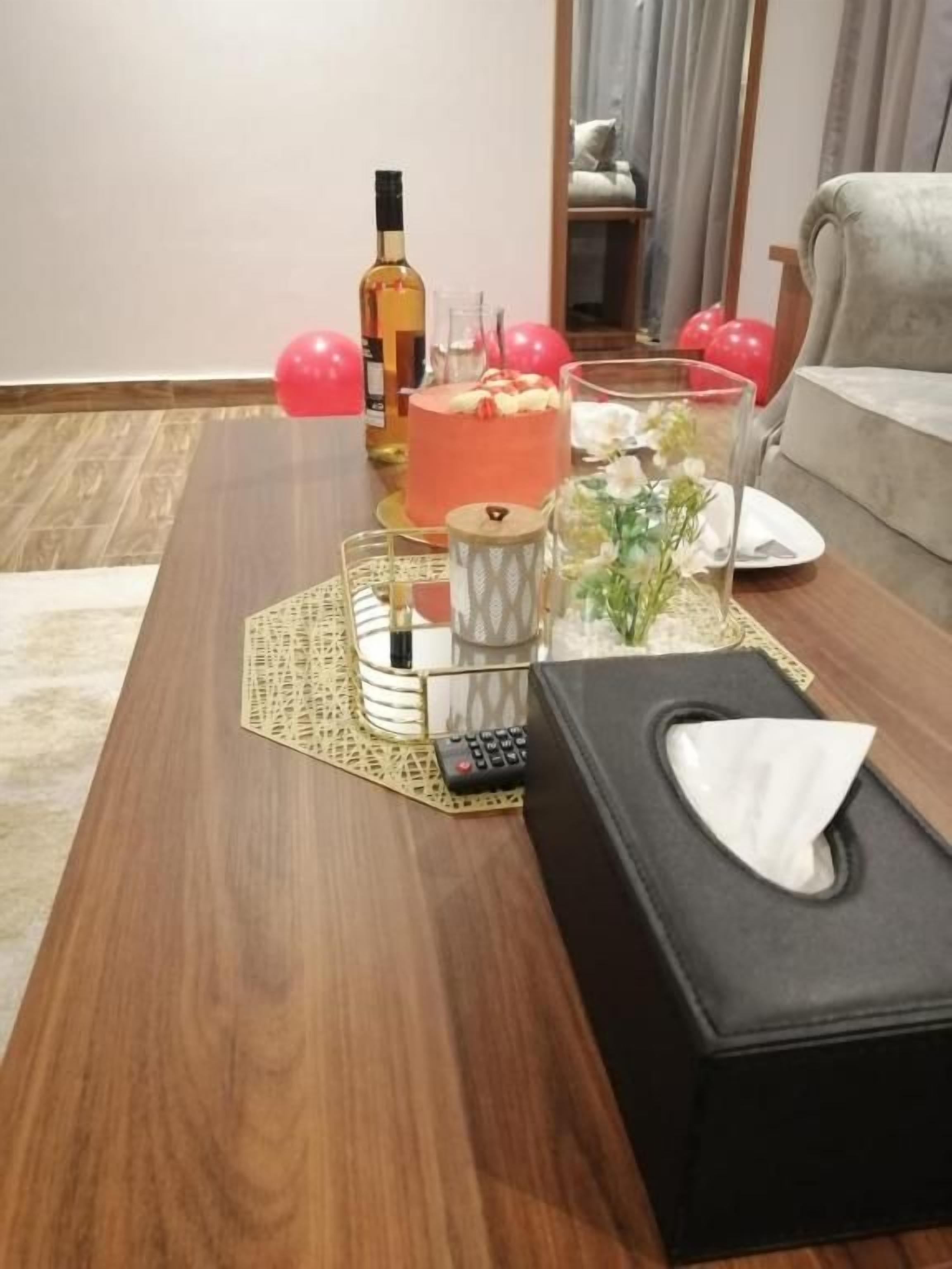 Una mesa de centro de madera con un control remoto, una caja de pañuelos, un vaso con flores, una botella y un pastel.