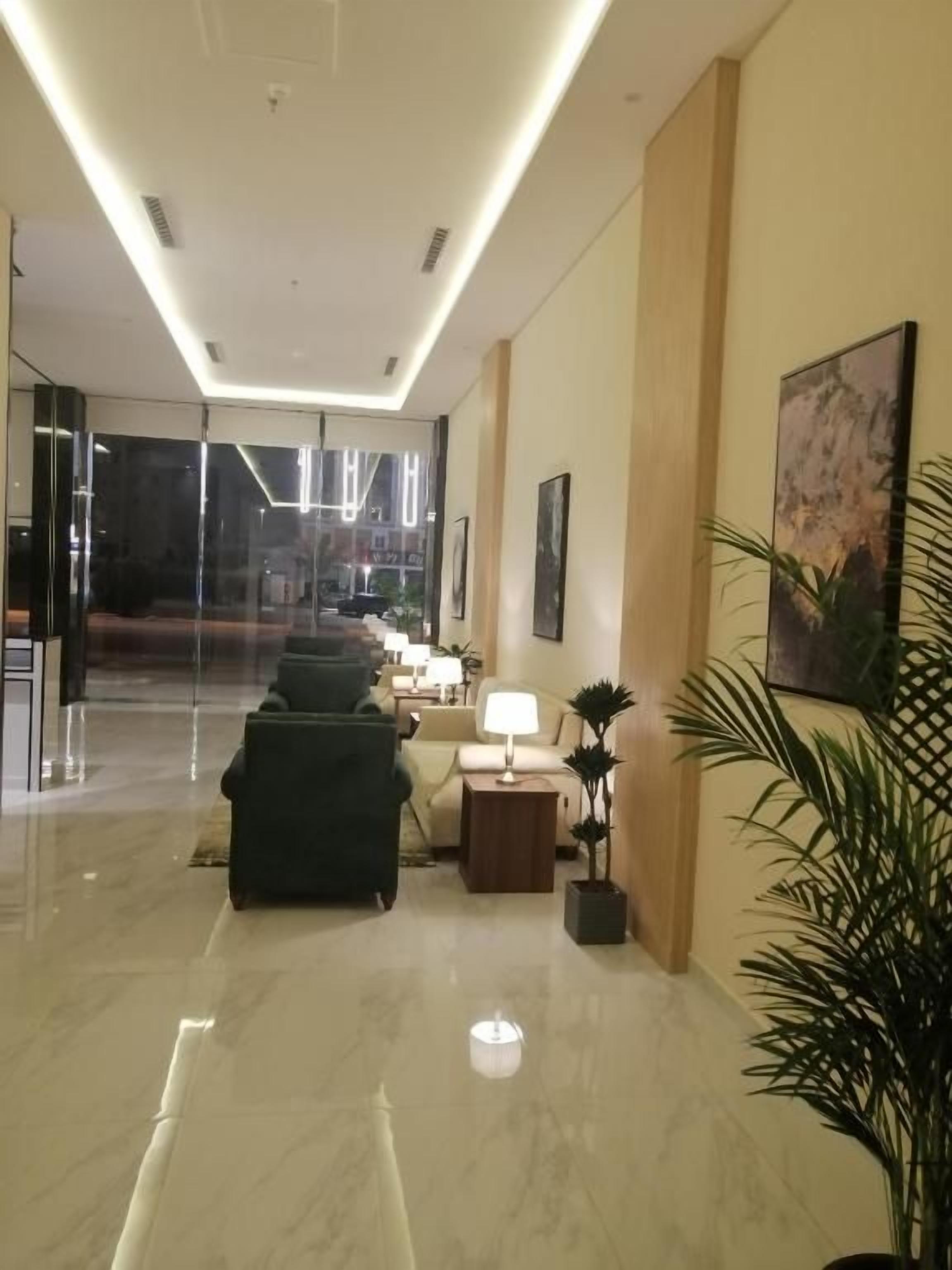 Vestíbulo de hotel con entrada por puerta de vidrio, zona de estar con lámpara y planta en maceta.