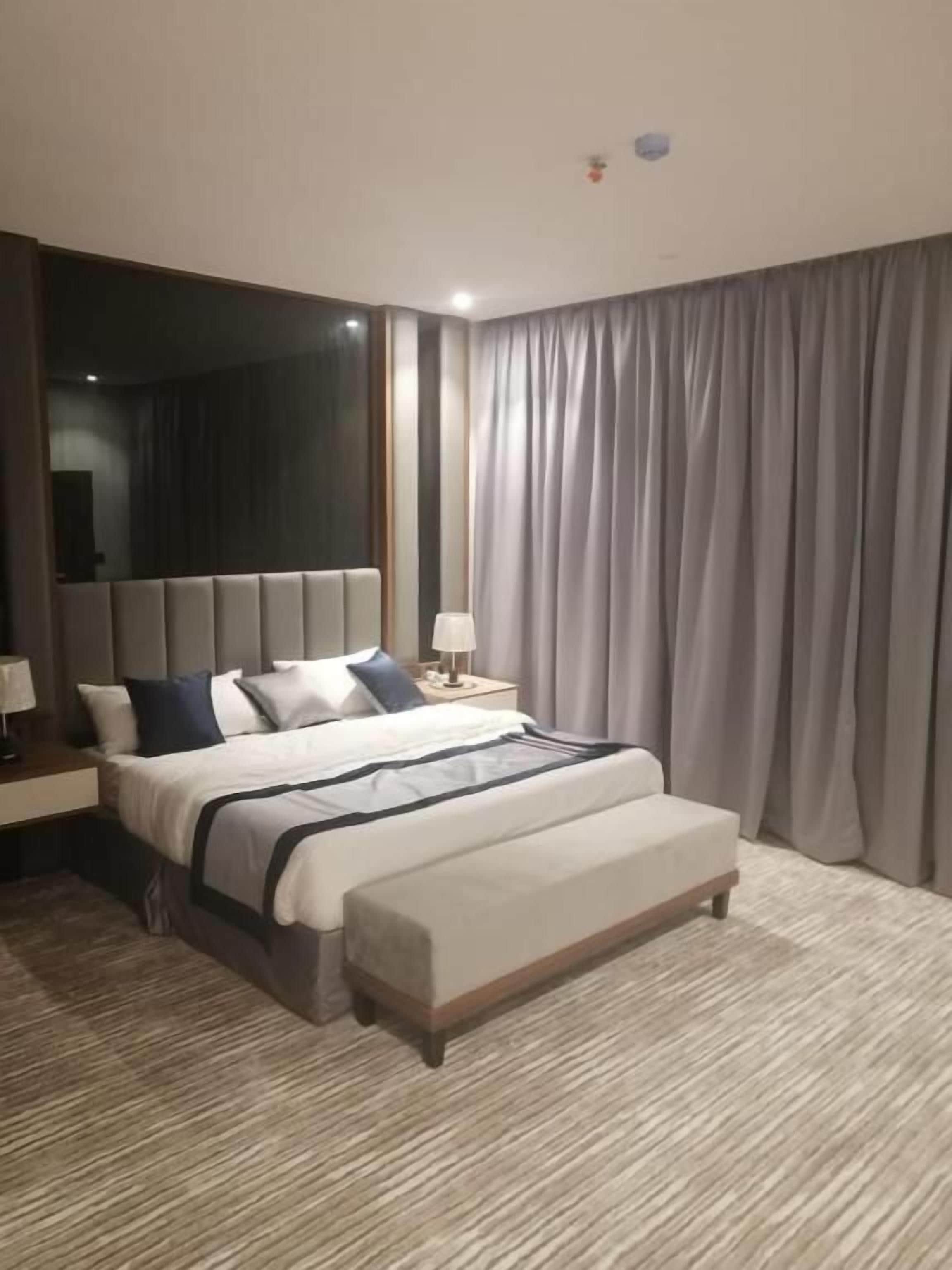 A hotel room with a large bed, a bedside table with a lamp, and a large window with curtains.