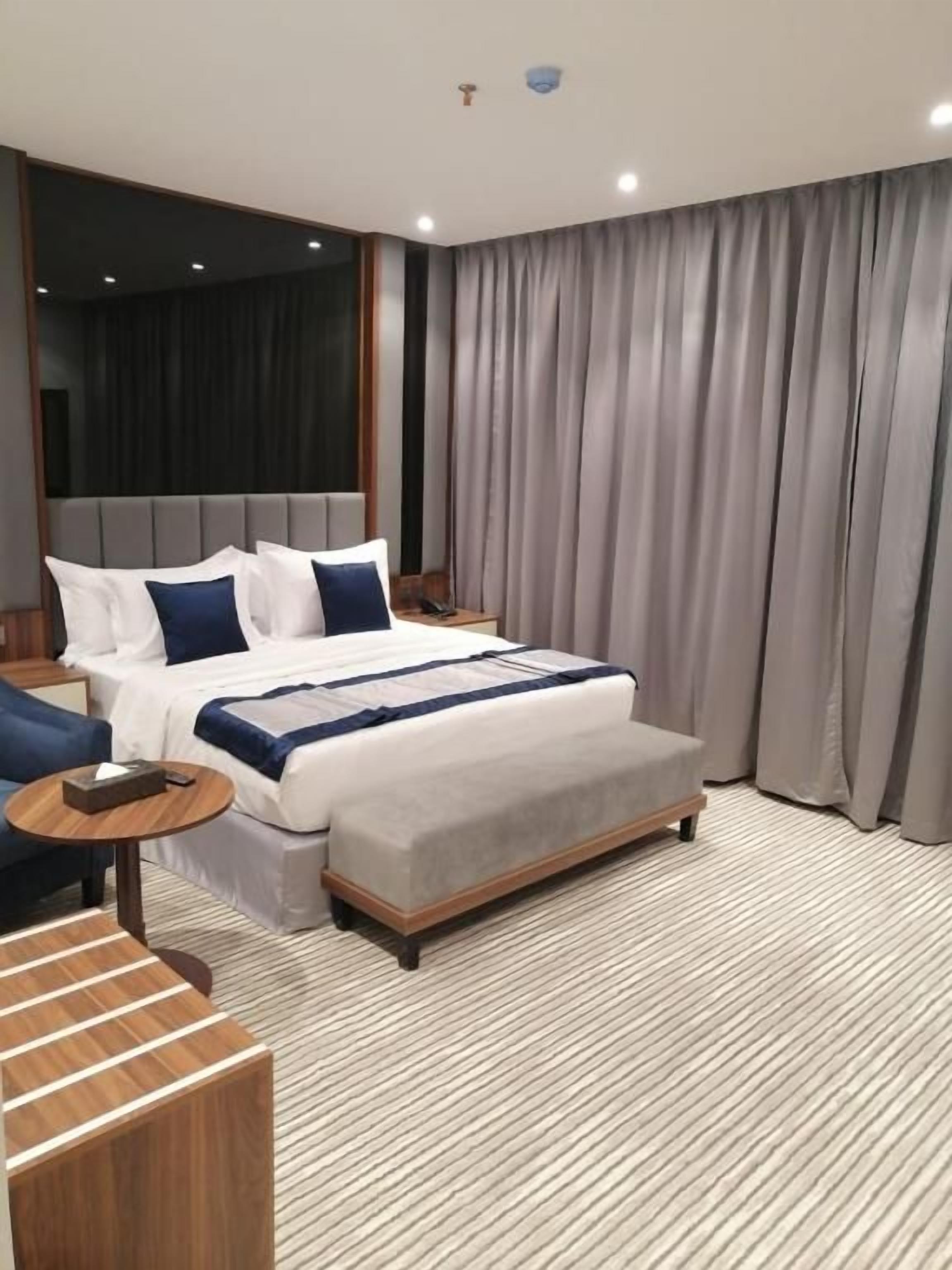 Una habitación de hotel moderna con una cama grande, una mesita de noche, una mesita auxiliar de madera y un ventanal con cortinas.