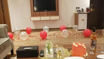 Una habitación con piso de madera, un televisor de pantalla plana montado en la pared, una pequeña kitchenette y una mesa de comedor con un pastel y vasos.