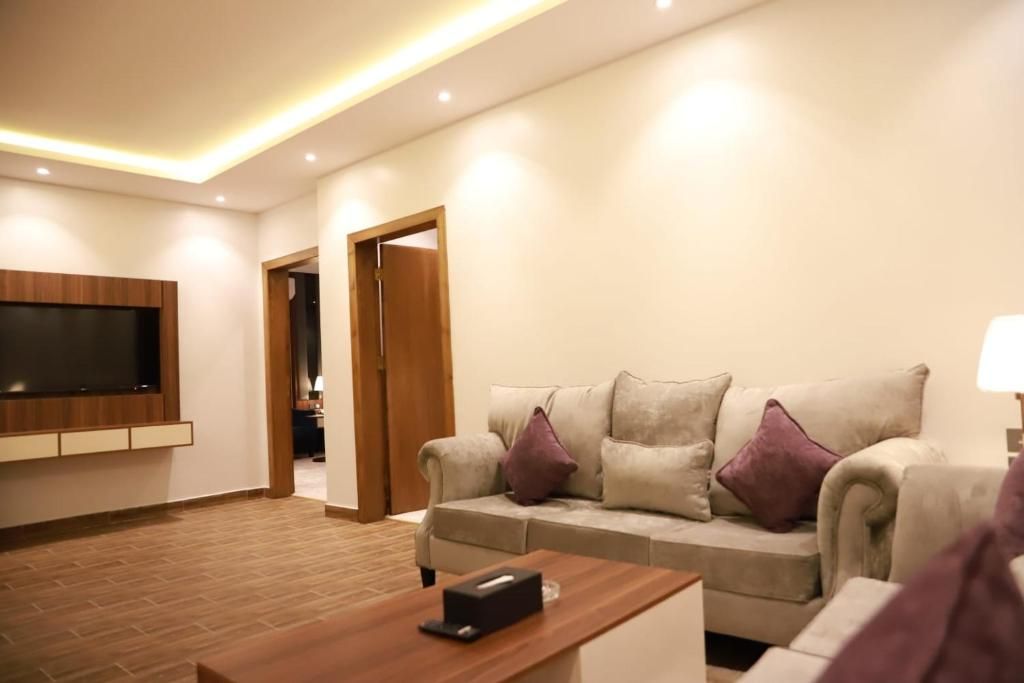 Una sala de estar moderna con un televisor de pantalla plana montado en la pared, un sofá color beige con cojines, una mesa de centro de madera y una puerta que da a otra habitación.