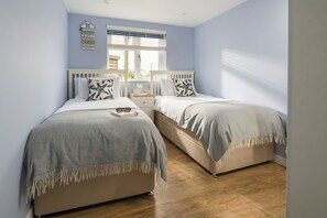 Ein Schlafzimmer mit zwei Betten, einem Fenster mit Jalousien, einem Nachttisch mit Tasse und Buch sowie einer Wanddekoration.
