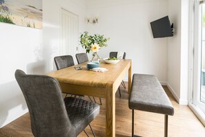 Ein Essbereich mit einem Holztisch, grau gepolsterten Stühlen, einer Bank, einer Vase mit Blumen und einem an der Wand montierten Fernseher.