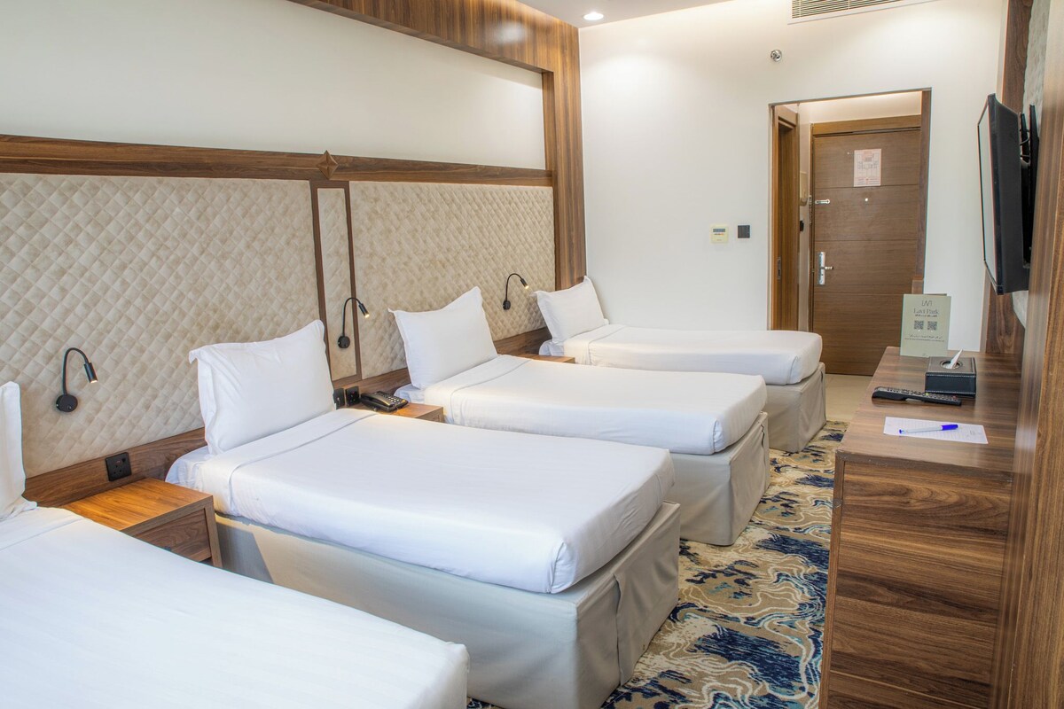 Ein Hotelzimmer mit drei Betten, einem Fernseher, einem Nachttisch mit Fernbedienung und einem gemusterten Teppich.