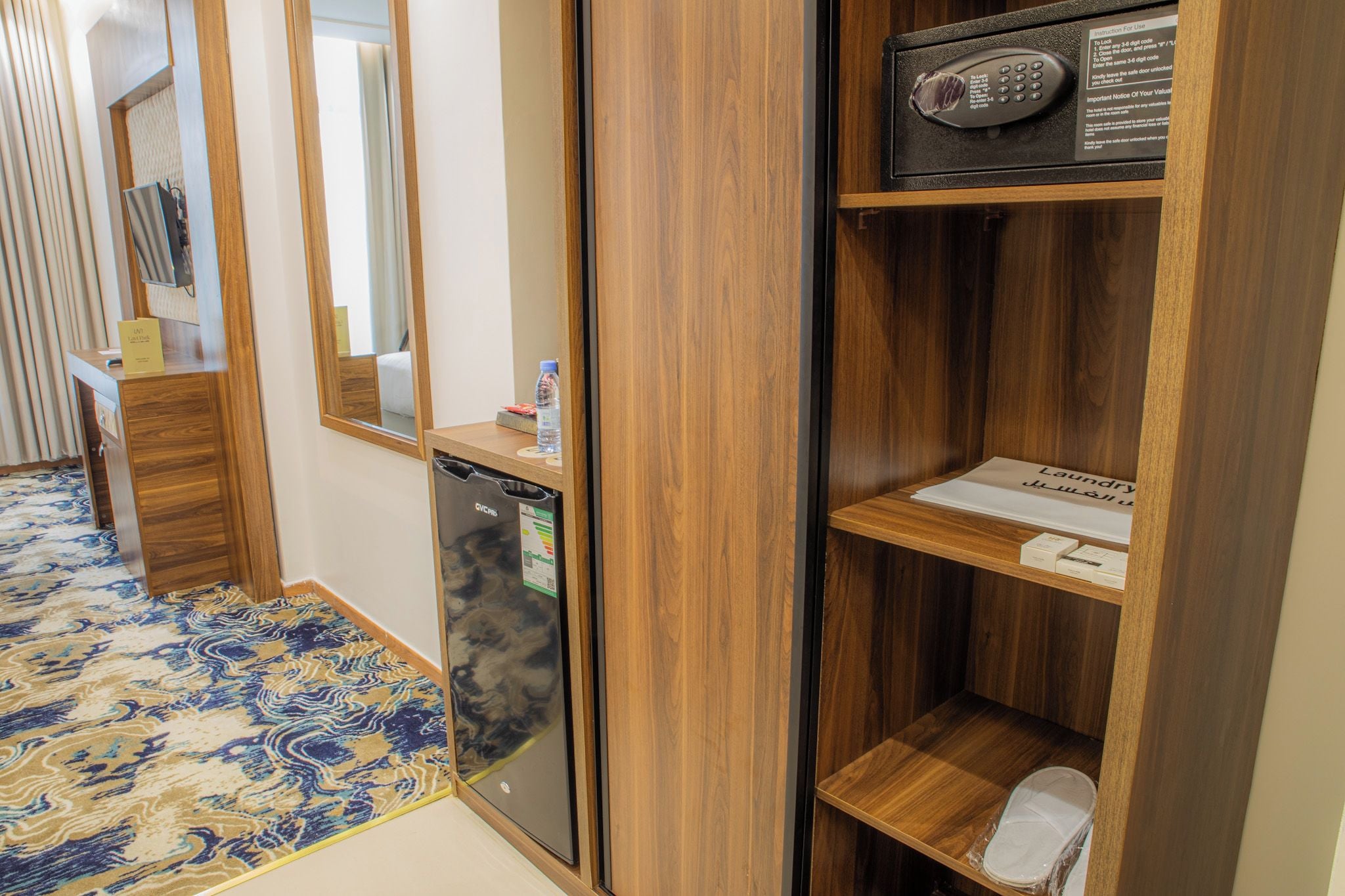A hotel room with a wooden wardrobe, a small desk with a water bottle, a trash bin, and a mirror.
