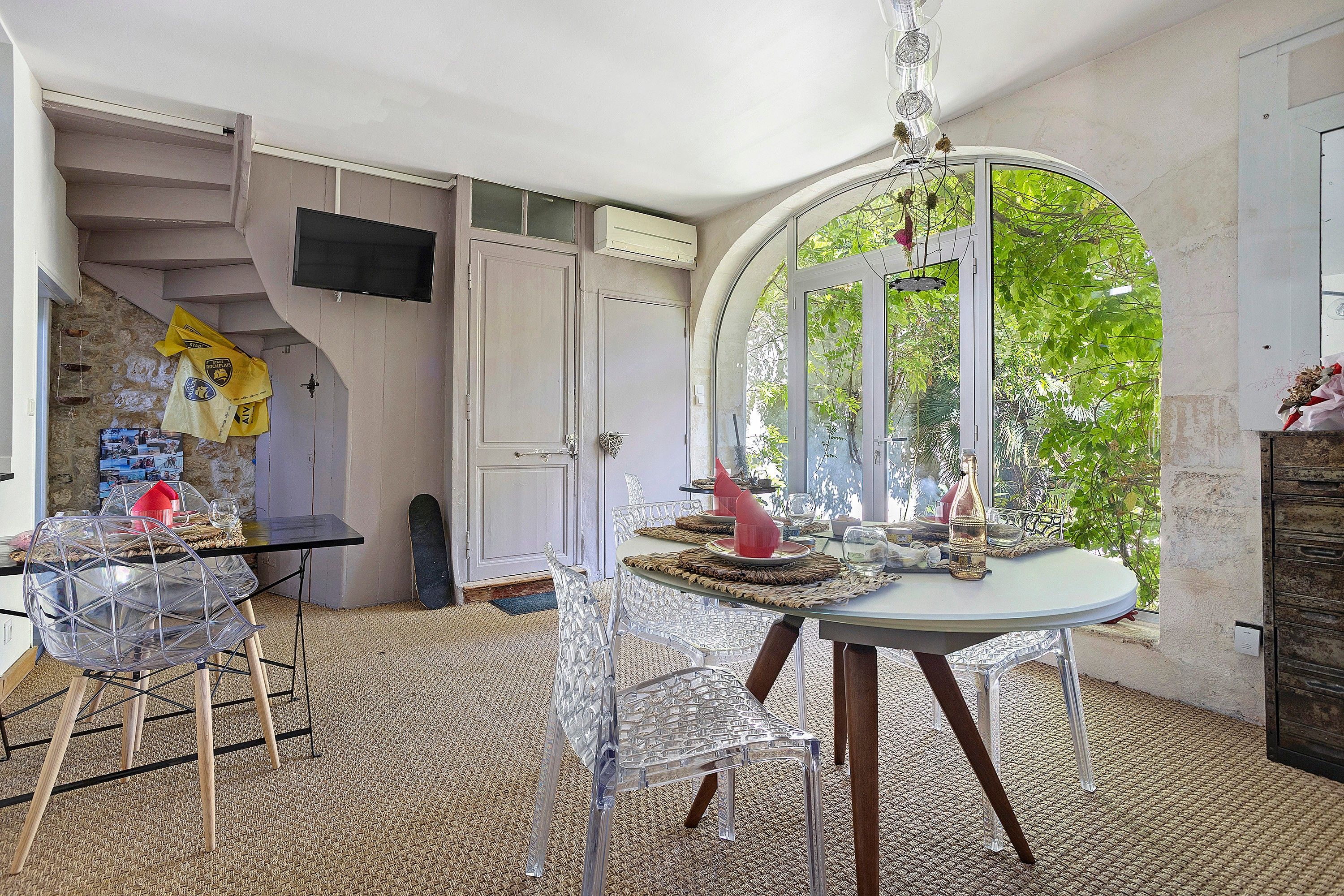 Ein Zimmer mit Esstisch, Stühlen, einem Fernseher und einer Tür zu einem Balkon mit Blick auf Grünflächen.