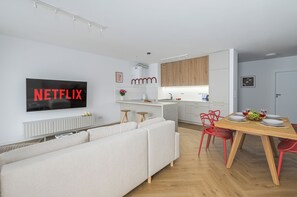 A modern living room with a white sofa, a dining table with red chairs, and a kitchen area with wooden cabinets. - Lumina 4-room Apart w/ Parking & Balcony (Warszawa)