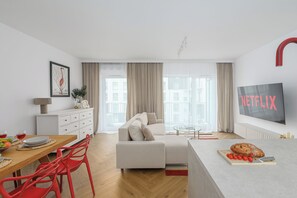 A modern living room with a sofa, a dining area with a table and chairs, a kitchen area with a fridge, and a large window with curtains. - Lumina 4-room Apart w/ Parking & Balcony (Warszawa)