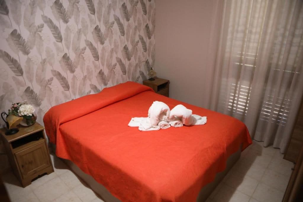 A hotel room with a bed, a nightstand, a small table with a vase, and a window with curtains.