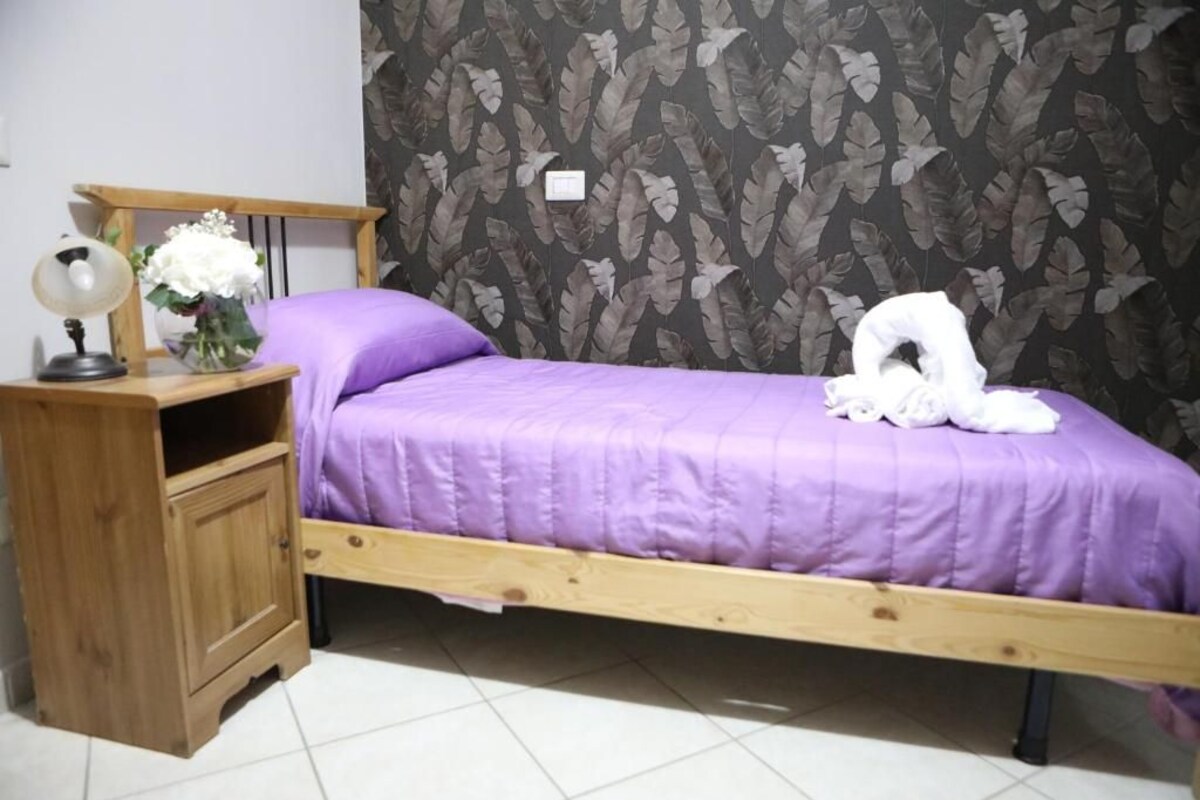 A single bed with a wooden frame, a purple bedspread, a wooden nightstand with a lamp, and a vase with flowers.