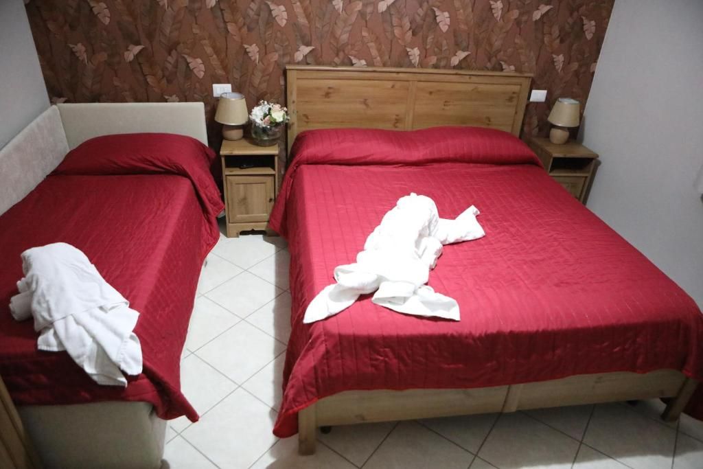 Una camera d'albergo con due letti singoli, ognuno con copriletto rosso e cuscini bianchi. Sono presenti due comodini con lampade e un vaso di fiori.