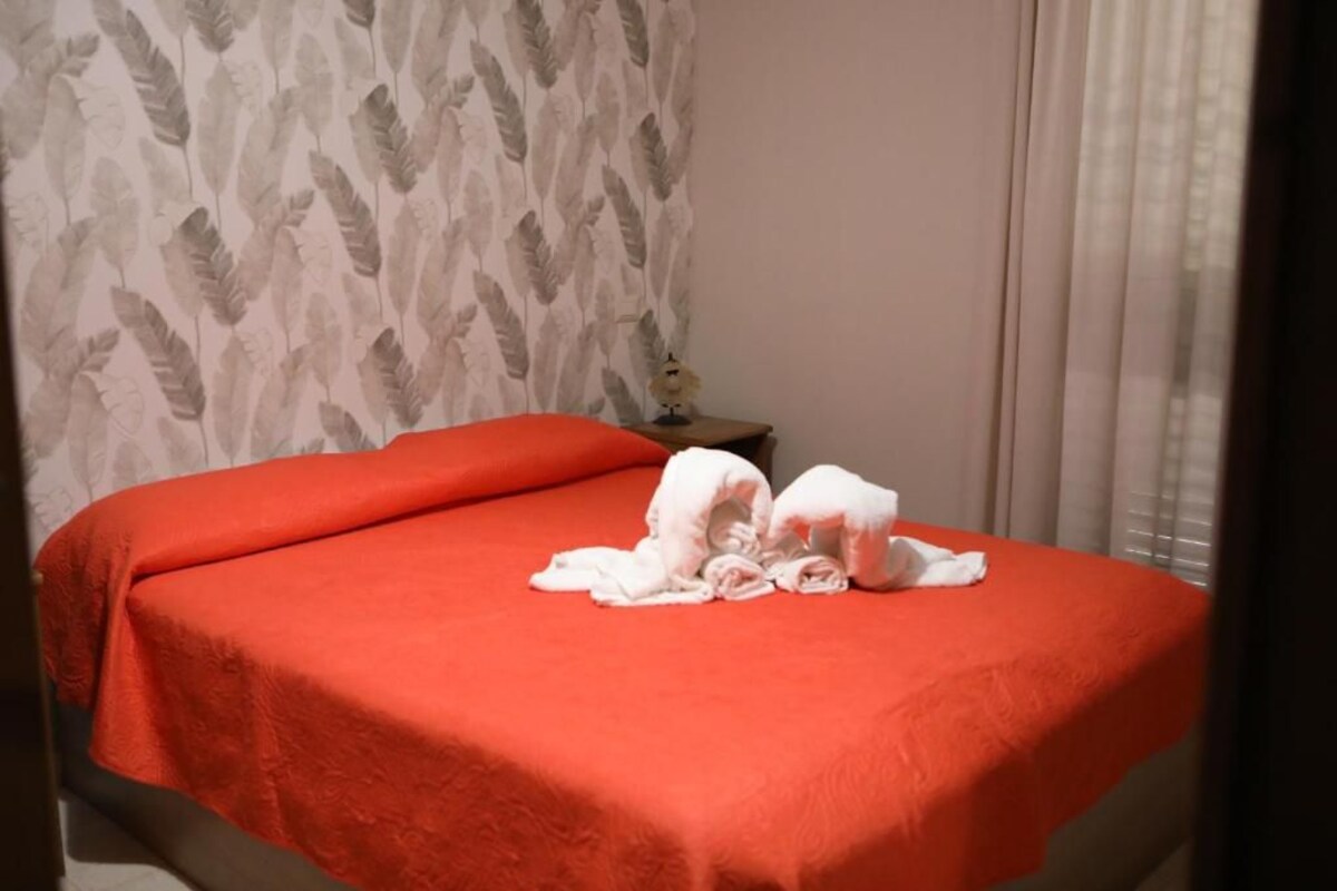 A hotel room with a bed covered in a red blanket, a white towel on the bed, and a nightstand with a lamp.