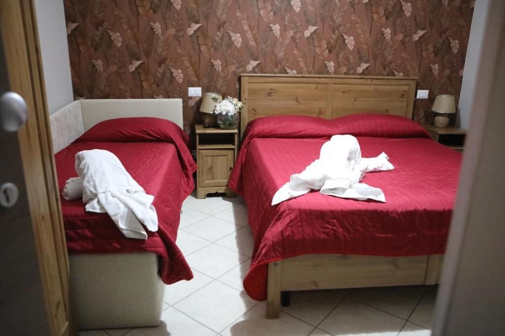 A room with two beds, each with a red bedspread and white pillows. There is a wooden nightstand with a lamp and a vase with flowers between the beds.
