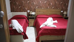 Une chambre avec deux lits, chacun recouvert d’une couverture rouge et agrémenté d’oreillers blancs. Une table de chevet en bois, sur laquelle se trouve une lampe, est placée entre les deux lits, ainsi qu’un vase avec des fleurs.