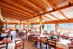 Uma sala de jantar espaçosa com vigas de madeira, mesas preparadas para refeições e grandes janelas que permitem a entrada de luz natural.