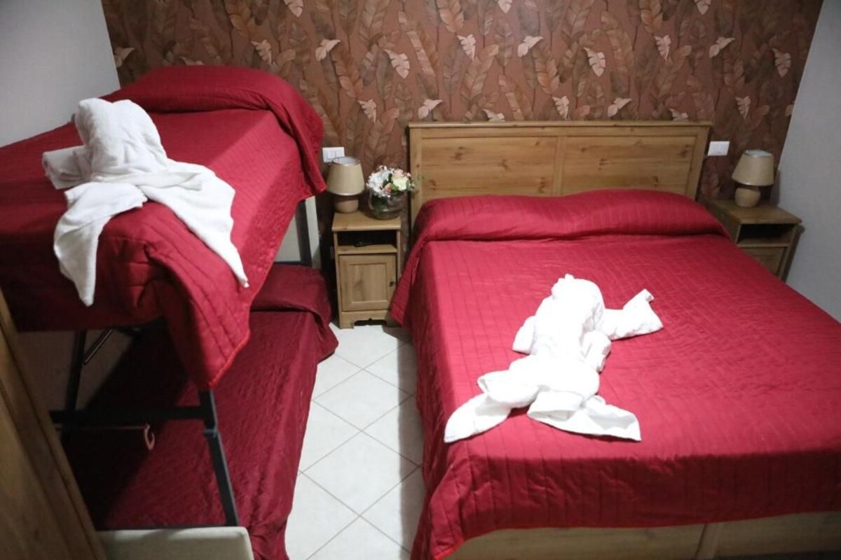 A hotel room with two beds, each with a red bedspread and white towels. There is a wooden headboard on the right bed, and a small table with a lamp and flowers on the left.