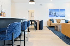Ein modernes Wohnzimmer mit einer Couch, Sesseln und einem Couchtisch. Es gibt eine Bar mit Hockern und einer Theke.