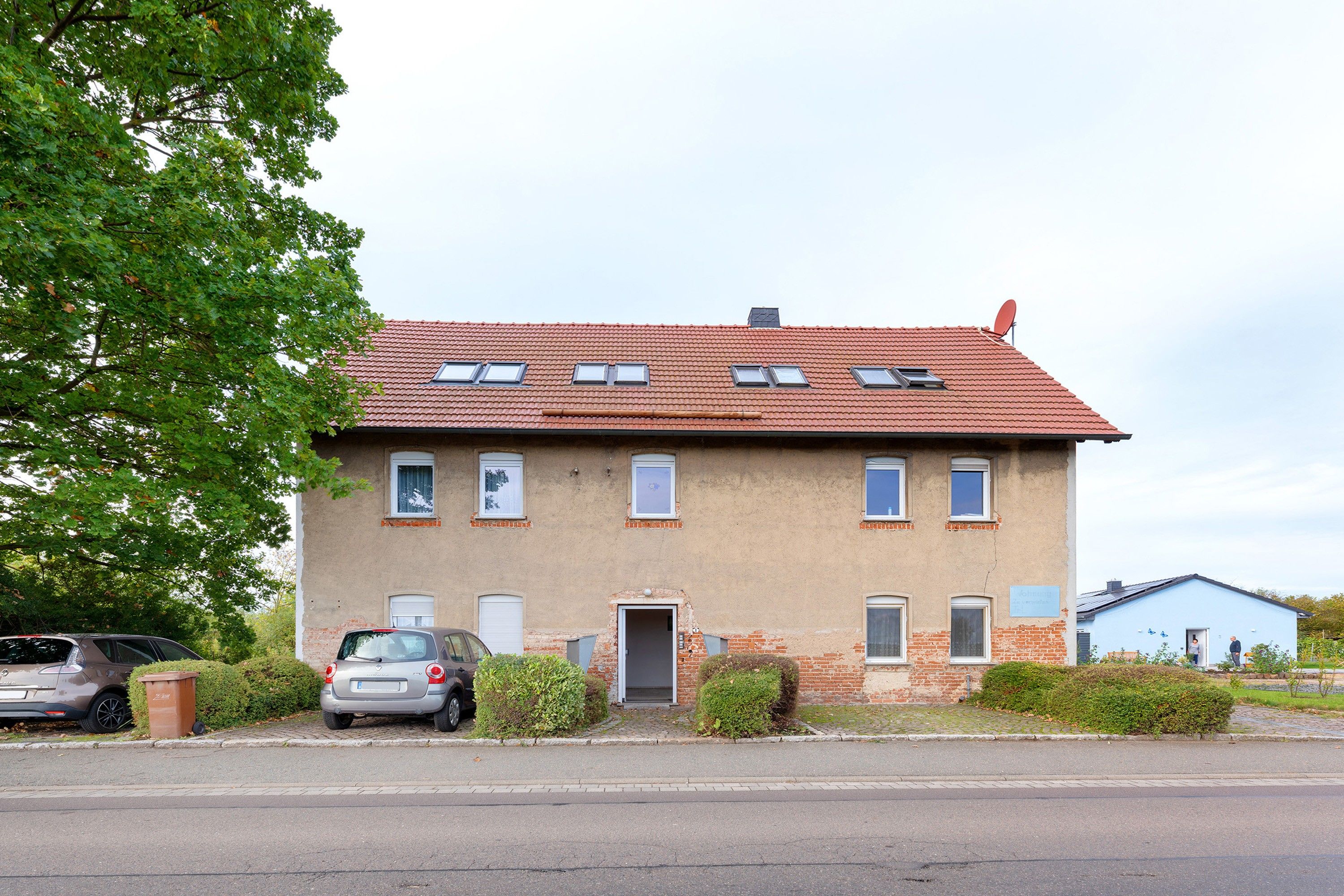 A two-story residential building with a red-tiled roof, multiple windows, and a parked car in front.