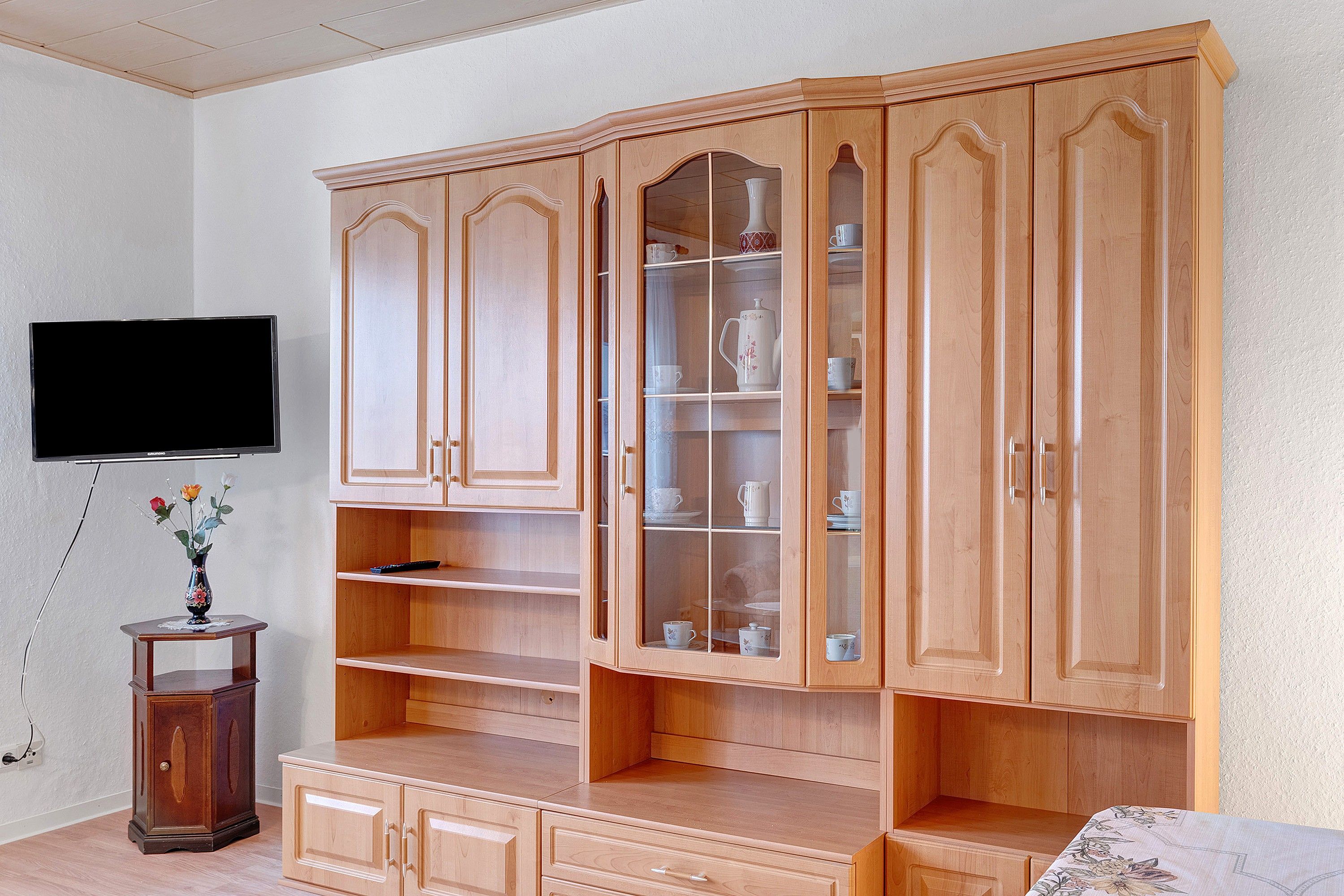 A wooden cabinet with multiple shelves and drawers, a television mounted on the wall, and a small table with a vase.