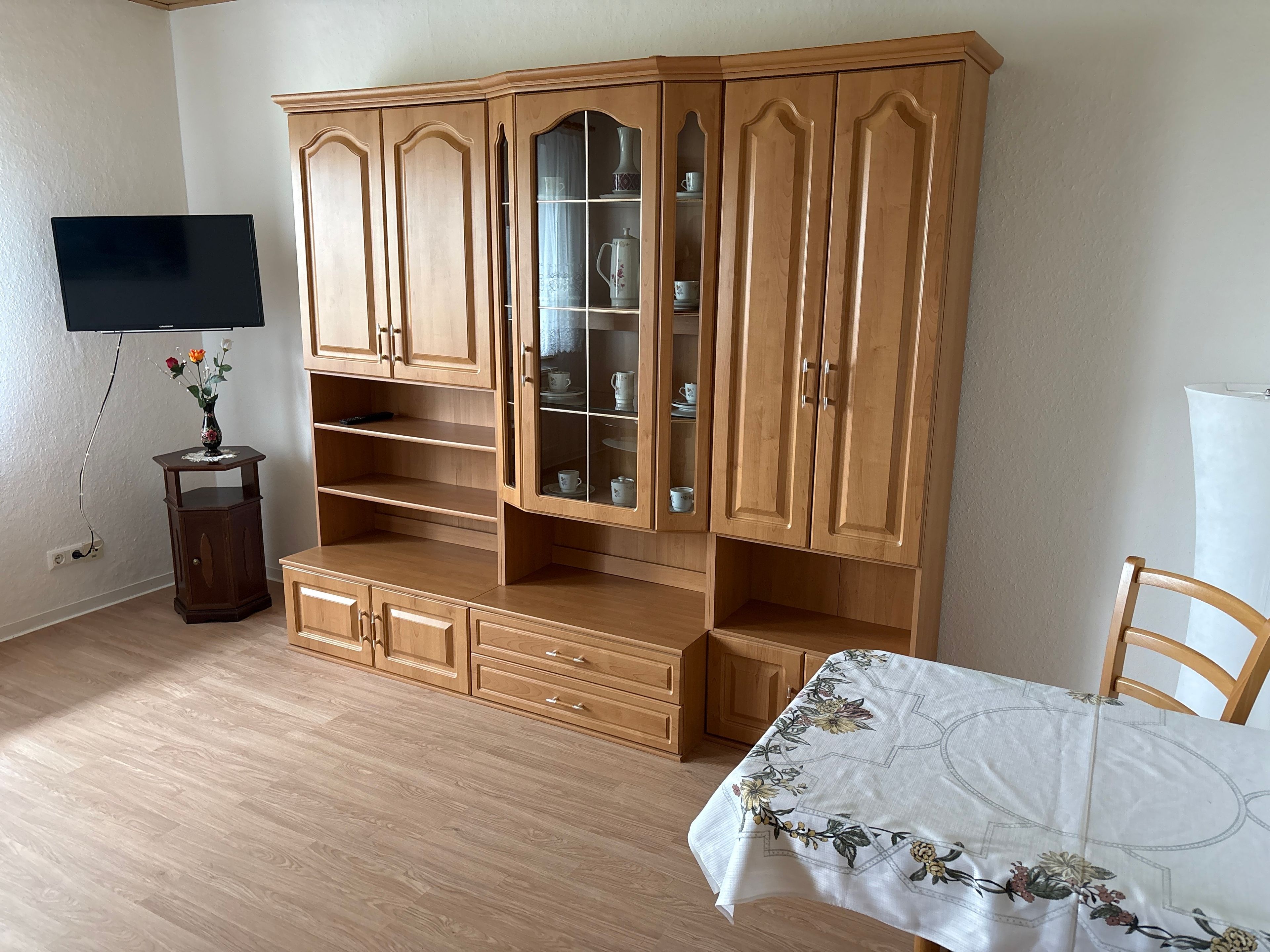 Una habitación con un armario de madera, un televisor montado en la pared y una mesa con mantel floreado.