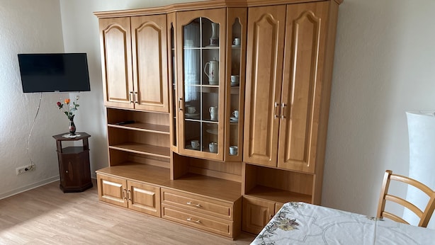 Una habitación con un armario de madera, un televisor montado en la pared y una mesa con mantel floreado.