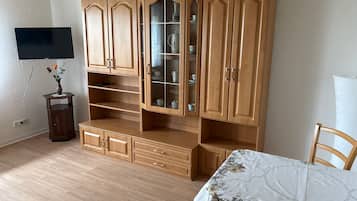 Una habitación con un armario de madera, un televisor montado en la pared y una mesa con mantel floreado.