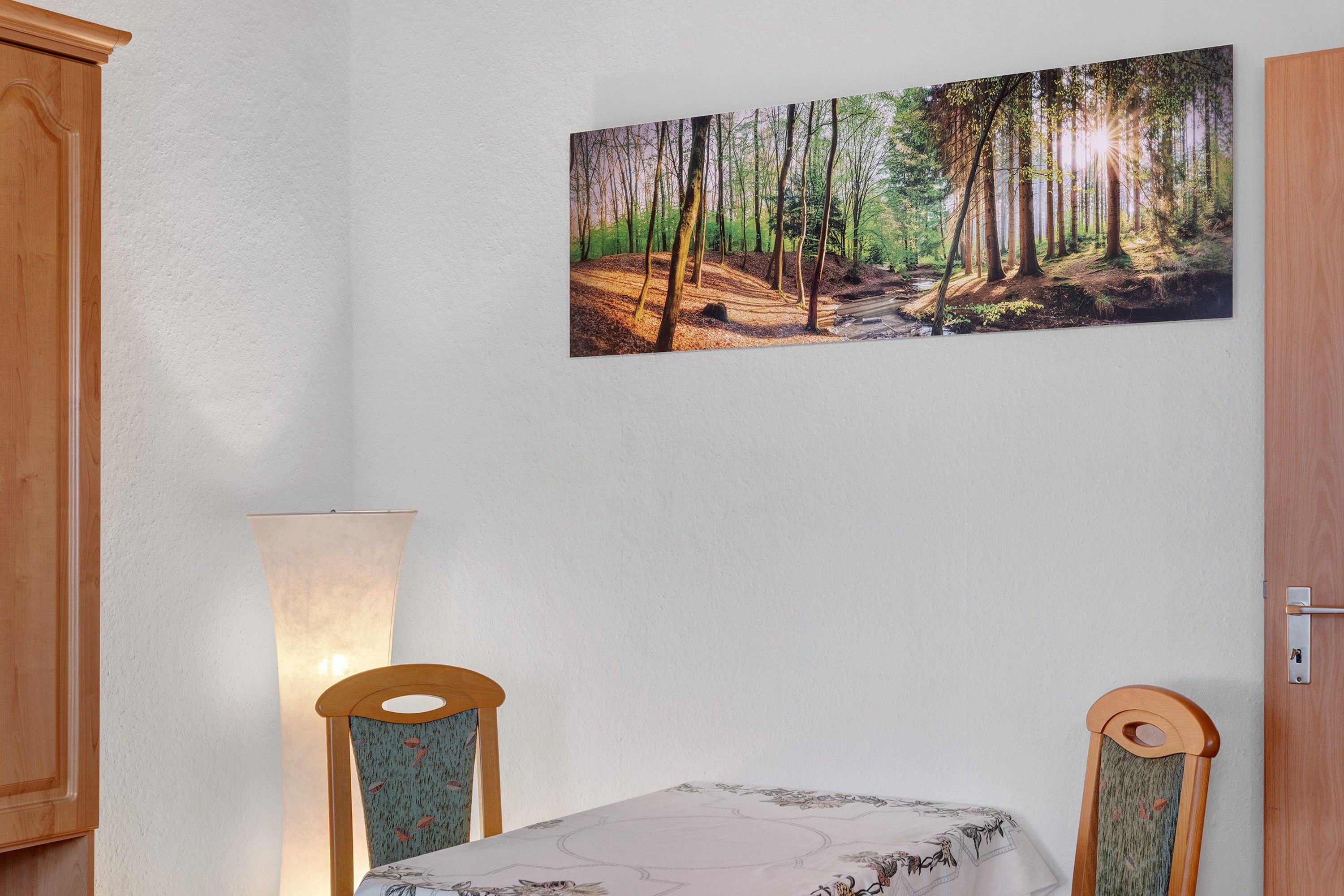A dining area with a table, chairs, a wall-mounted painting of a forest, and a lamp.