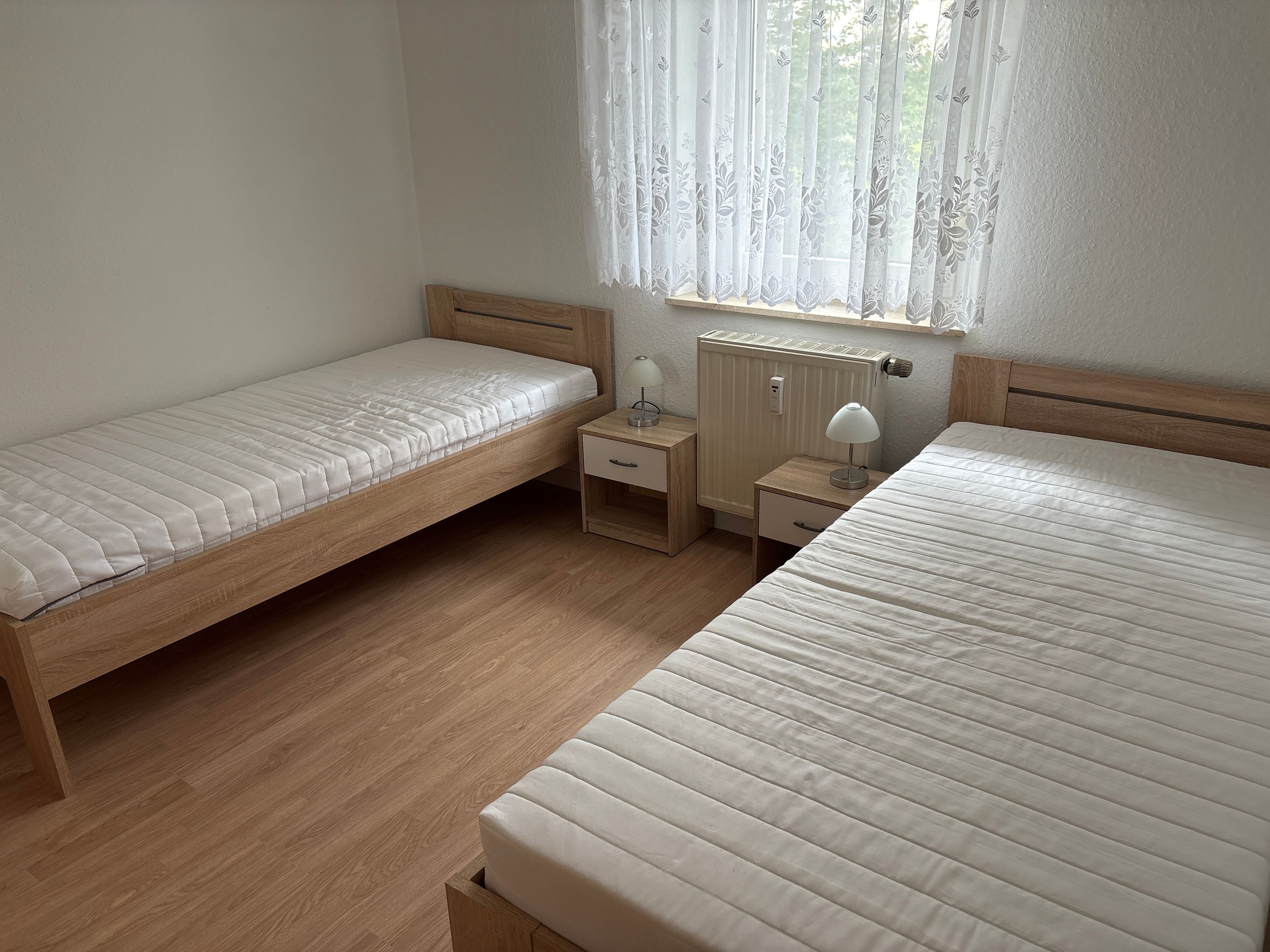 Un dormitorio con dos camas individuales, una mesita de noche con lámpara y una ventana con cortinas traslúcidas.