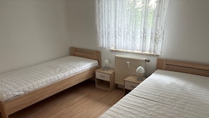 Un dormitorio con dos camas individuales, una mesita de noche con lámpara y una ventana con cortinas traslúcidas.