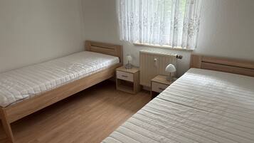 Un dormitorio con dos camas individuales, una mesita de noche con lámpara y una ventana con cortinas traslúcidas.