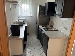 Eine kleine Küche mit Kaffeemaschine, Kühlschrank, Spüle und einem Fenster mit Vorhängen.