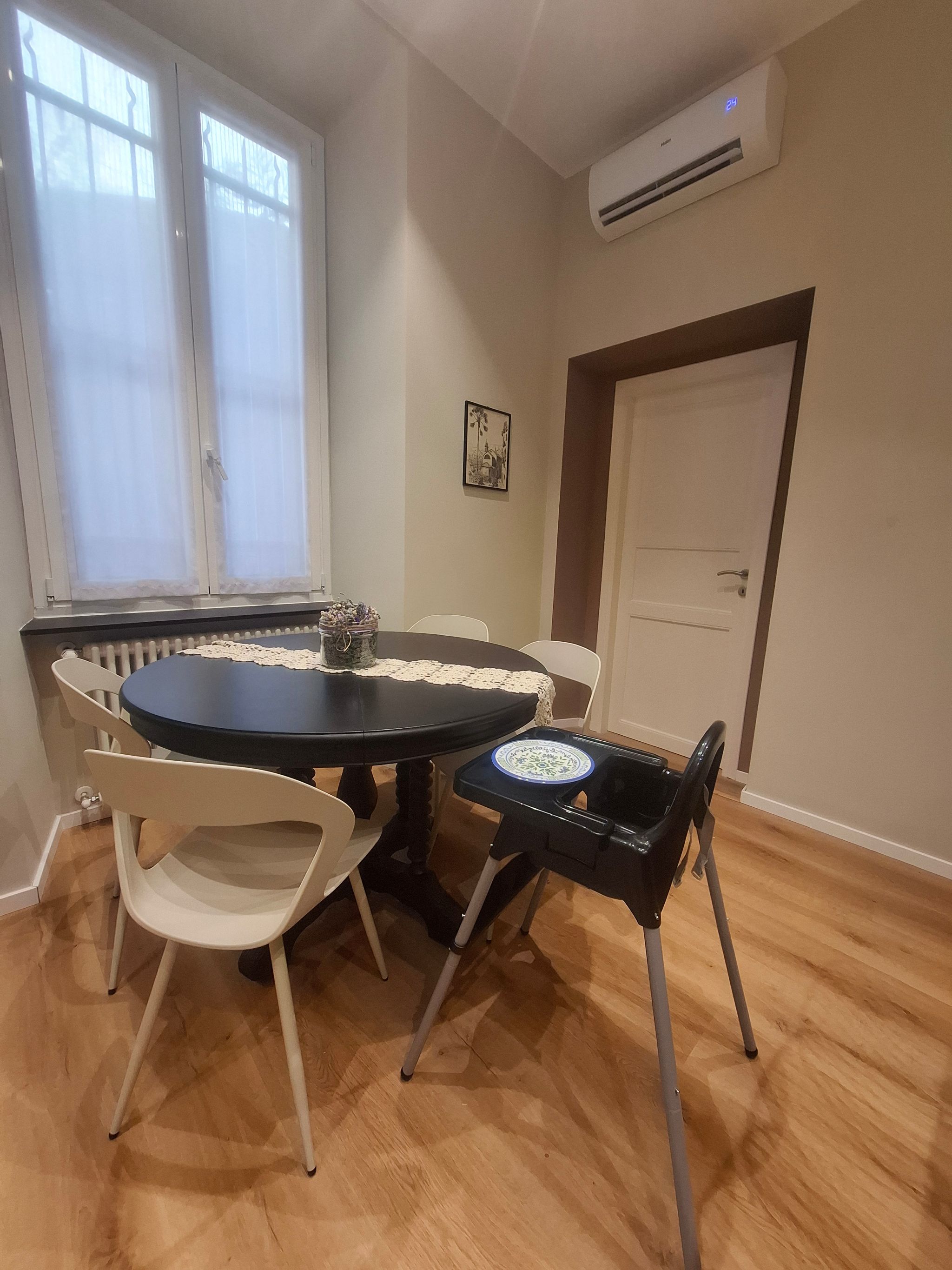 A dining area with a round table, white chairs, a black chair with a tray, and an air conditioning unit on the wall.