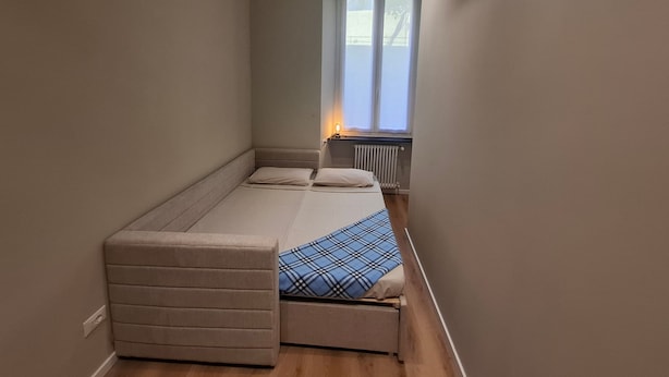 Een kleine kamer met een bed, een bankje, een radiator en een raam.