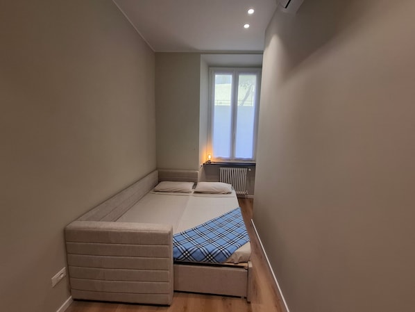 Una habitación pequeña con una cama, un banco, un radiador y una ventana.