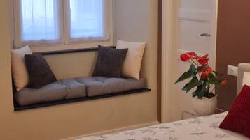 A room with a built-in bench against the wall, a potted plant with red flowers, and a bed with a white headboard.