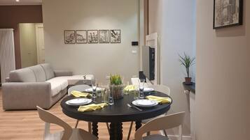 Un comedor con una mesa redonda, sillas blancas y un mantel negro. Hay cuadros en la pared y una planta en maceta al lado.
