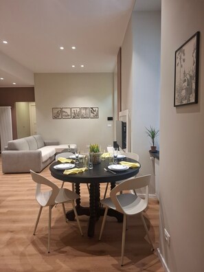 Un coin repas avec une table ronde, des chaises blanches et une nappe noire. Des tableaux encadrés ornent le mur, et une plante en pot est posée sur le côté.