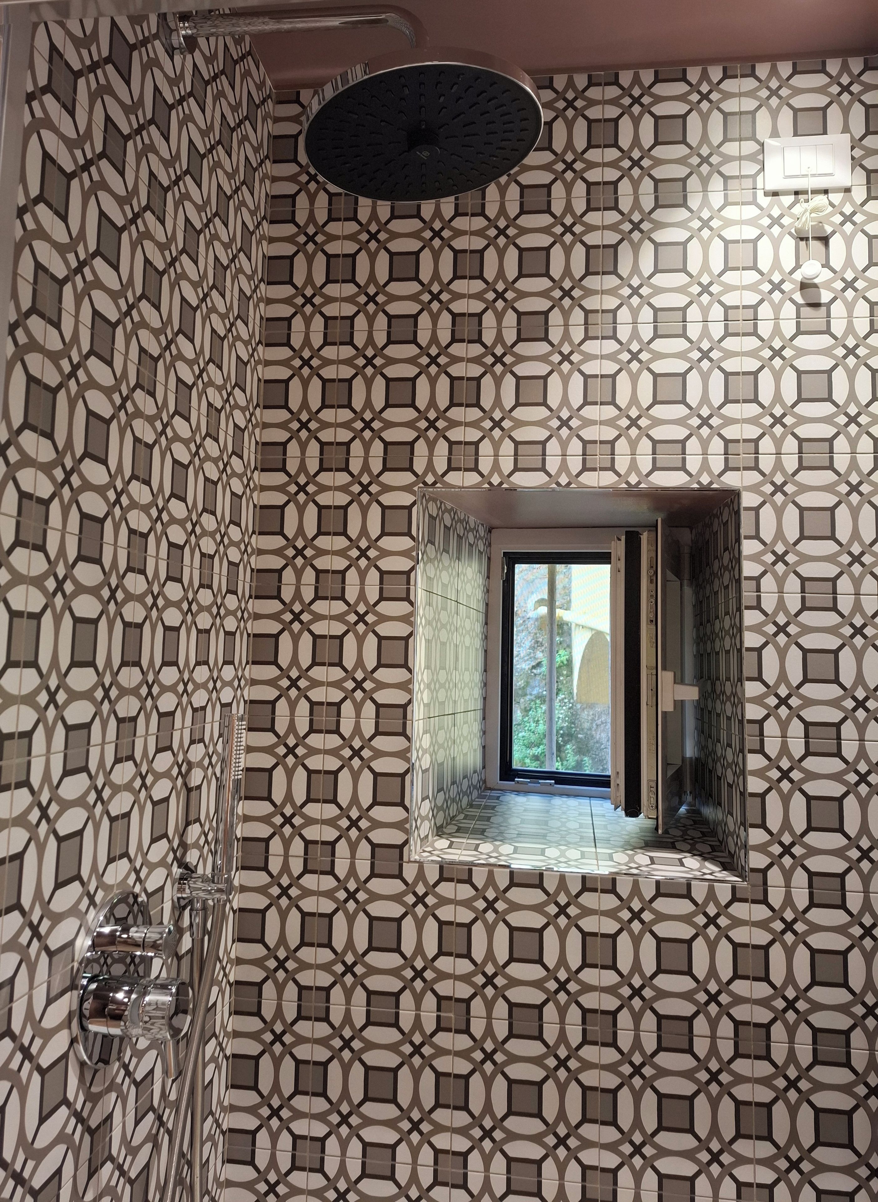 Un baño con diseño de baldosas geométricas, una ducha con grifería moderna y una ventana con vista a follaje.