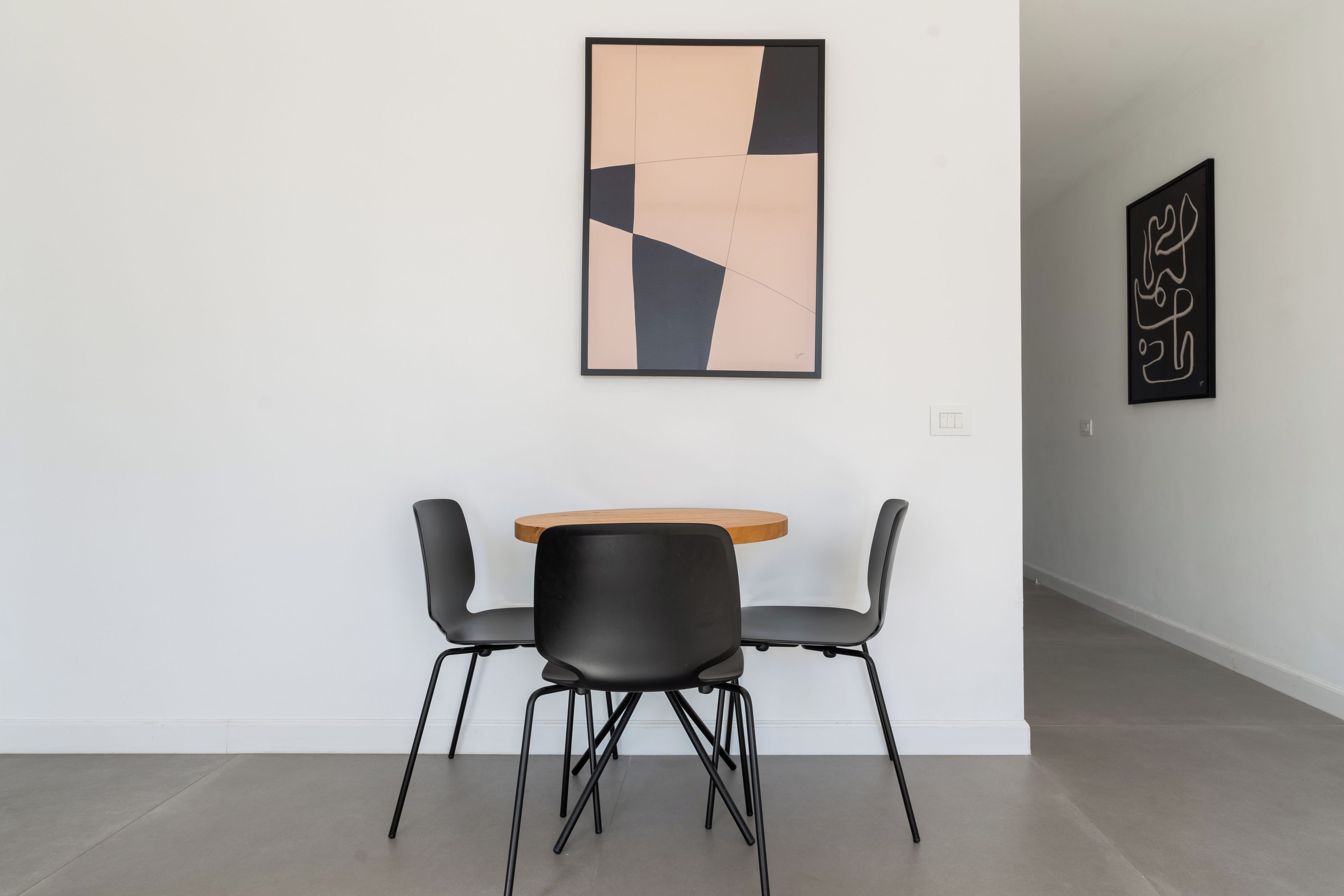 A minimalist room with a round wooden table and two black chairs, a geometric abstract painting on the wall, and a plain white wall.