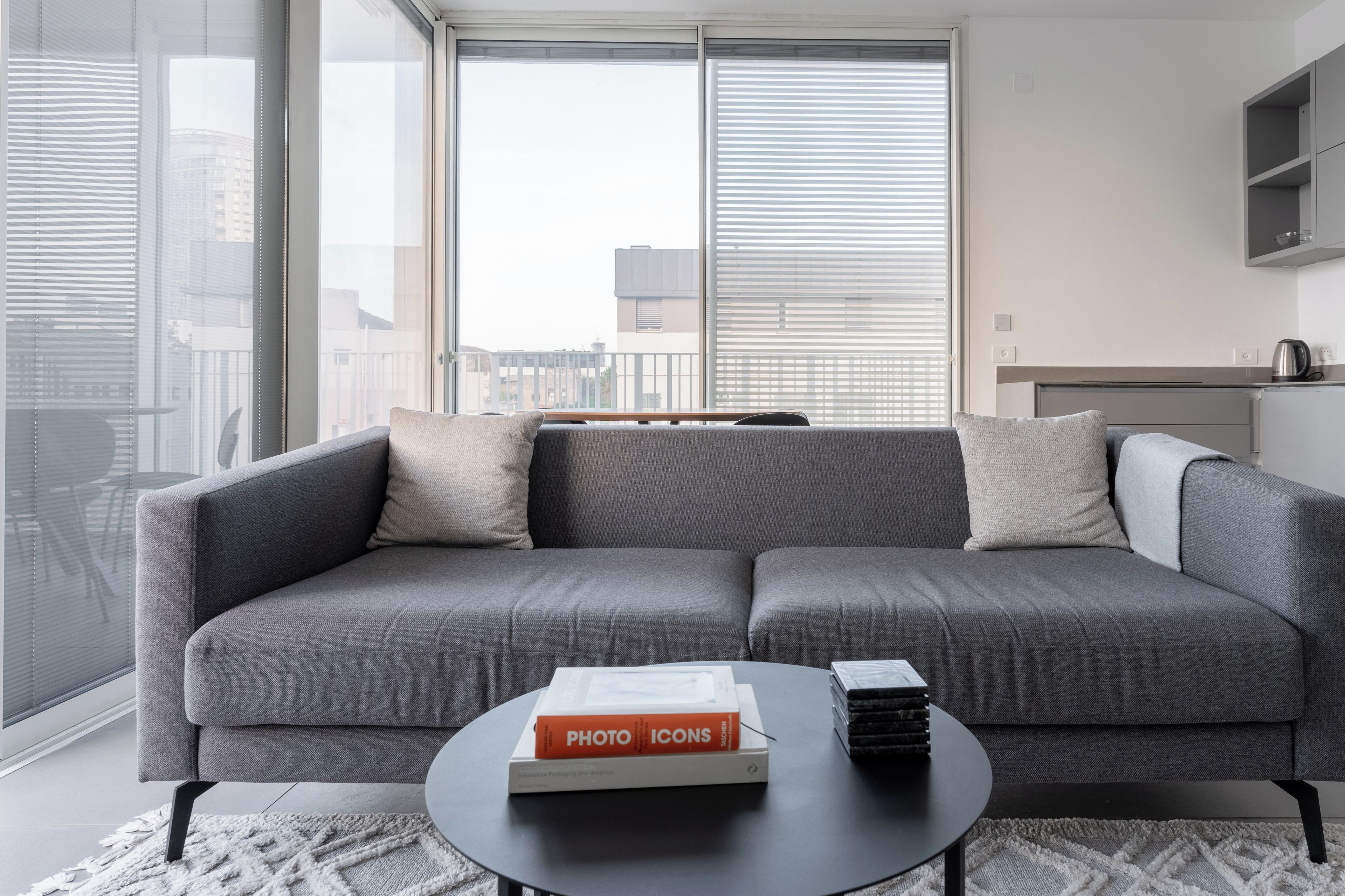 A modern living room with a grey sectional sofa, a coffee table with a book and a stack of magazines, and large windows with blinds.
