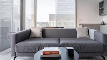 A modern living room with a grey sectional sofa, a coffee table with a book and a stack of magazines, and large windows with blinds.