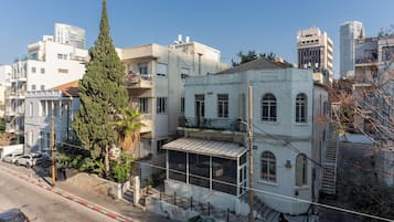 A multi-story building with a balcony, a tall tree in front, and a street with parked cars.
