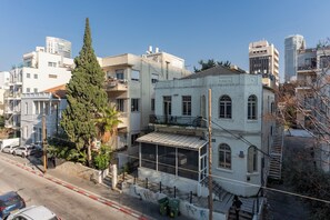 Un edifici de diversos pisos amb un balcó, un arbre alt al davant i un carrer amb cotxes aparcats.