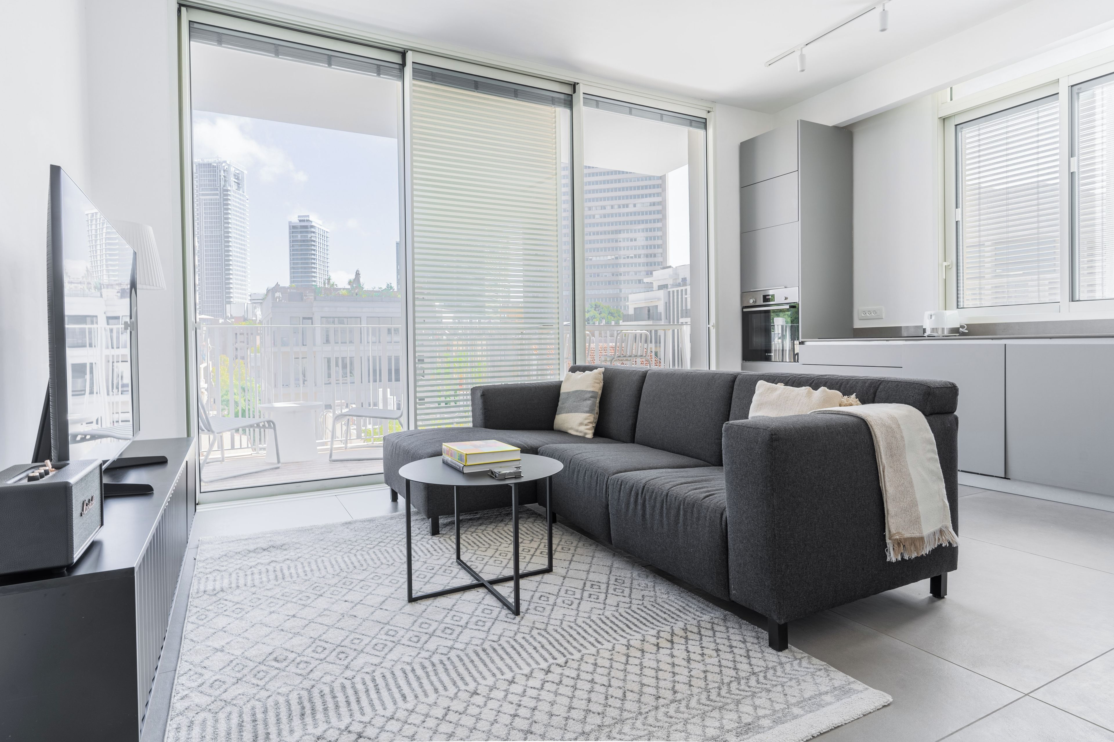 Un salon moderne avec un canapé gris, une petite table basse ronde et une vue sur la ville grâce à de grandes fenêtres.