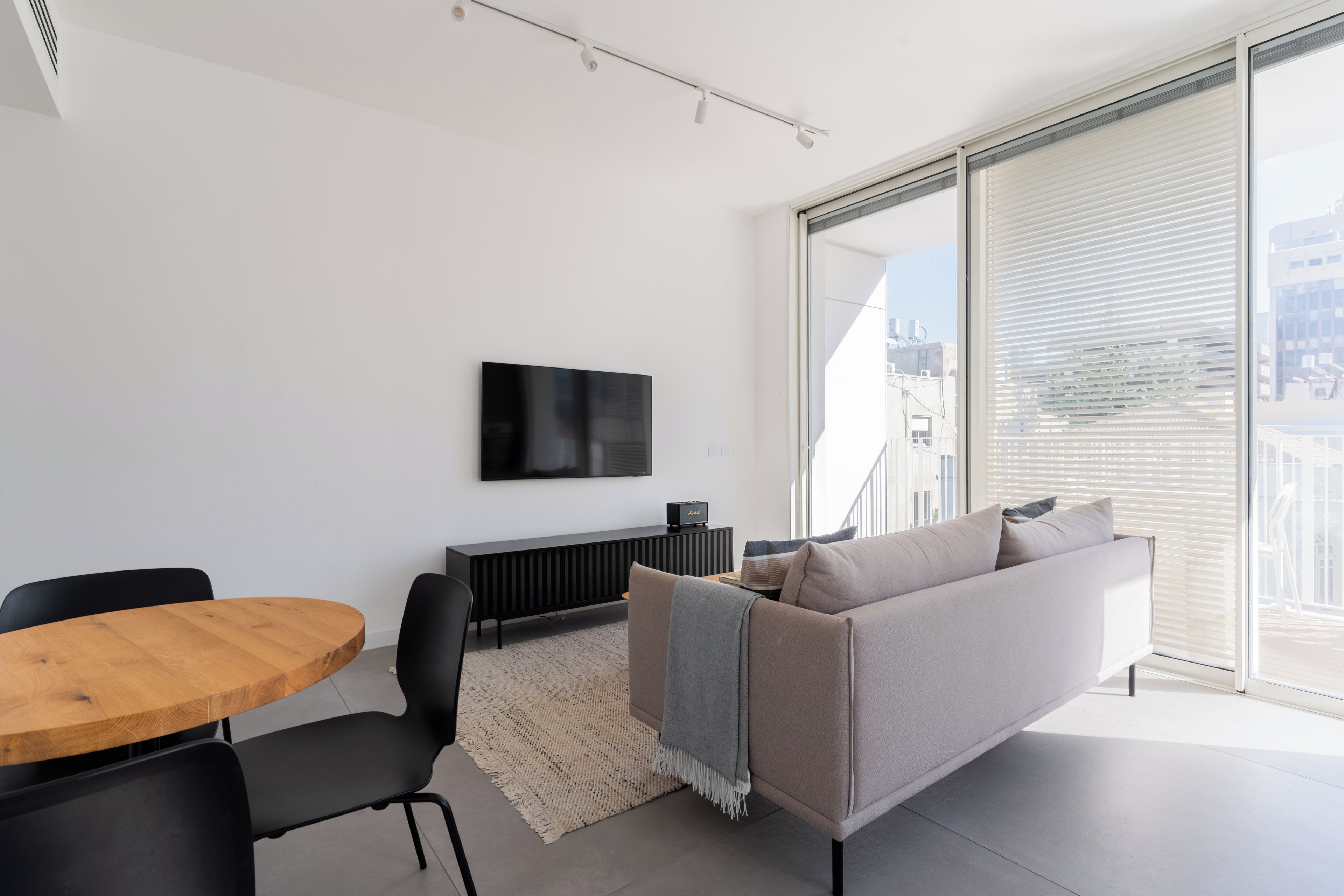 A modern living room with a flat-screen TV mounted on the wall, a round wooden table, a black sofa, and a large window with blinds.