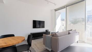 A modern living room with a flat-screen TV mounted on the wall, a round wooden table, a black sofa, and a large window with blinds.